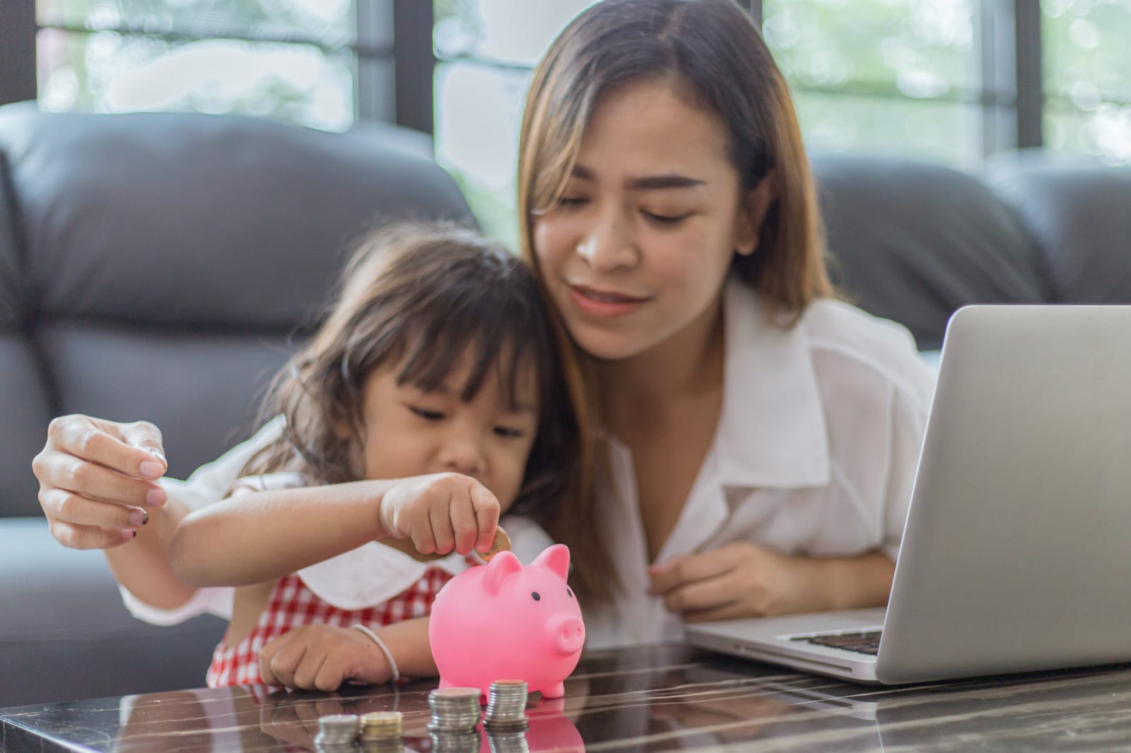 母親と小さな娘がノートパソコンのそばで貯金箱に硬貨を入れる様子